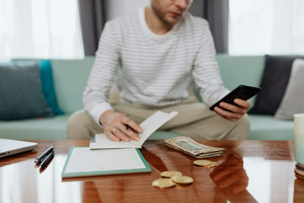 Adult man sits on sofa managing finances with cash, smartphone, and notebook.